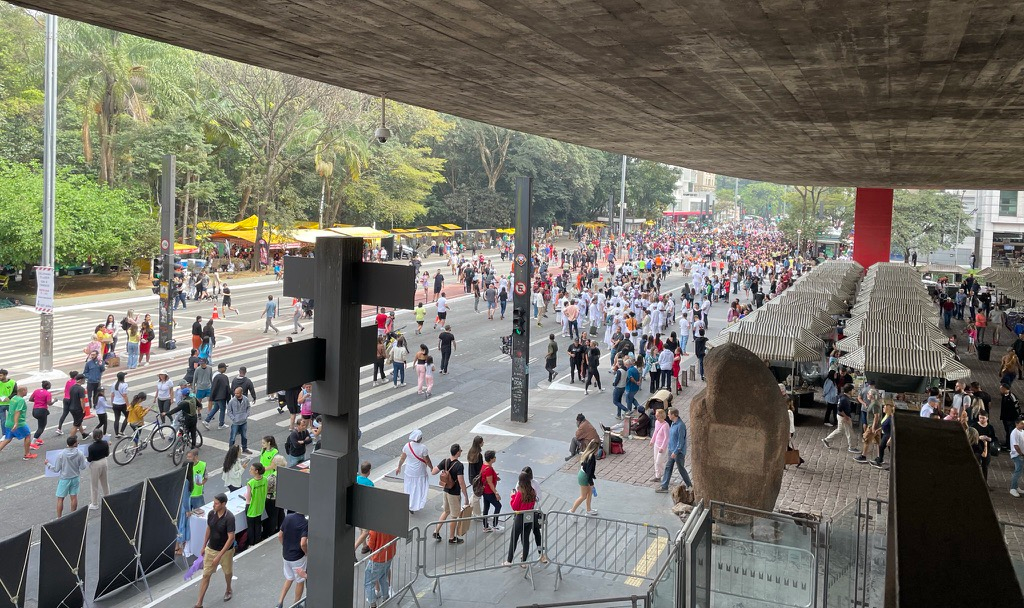 Picture taken from below the MASP museum, in São Paulo. The sidewalk and the street are filled with all kinds of people, and on the other side of the street, there are street food stands and a park.