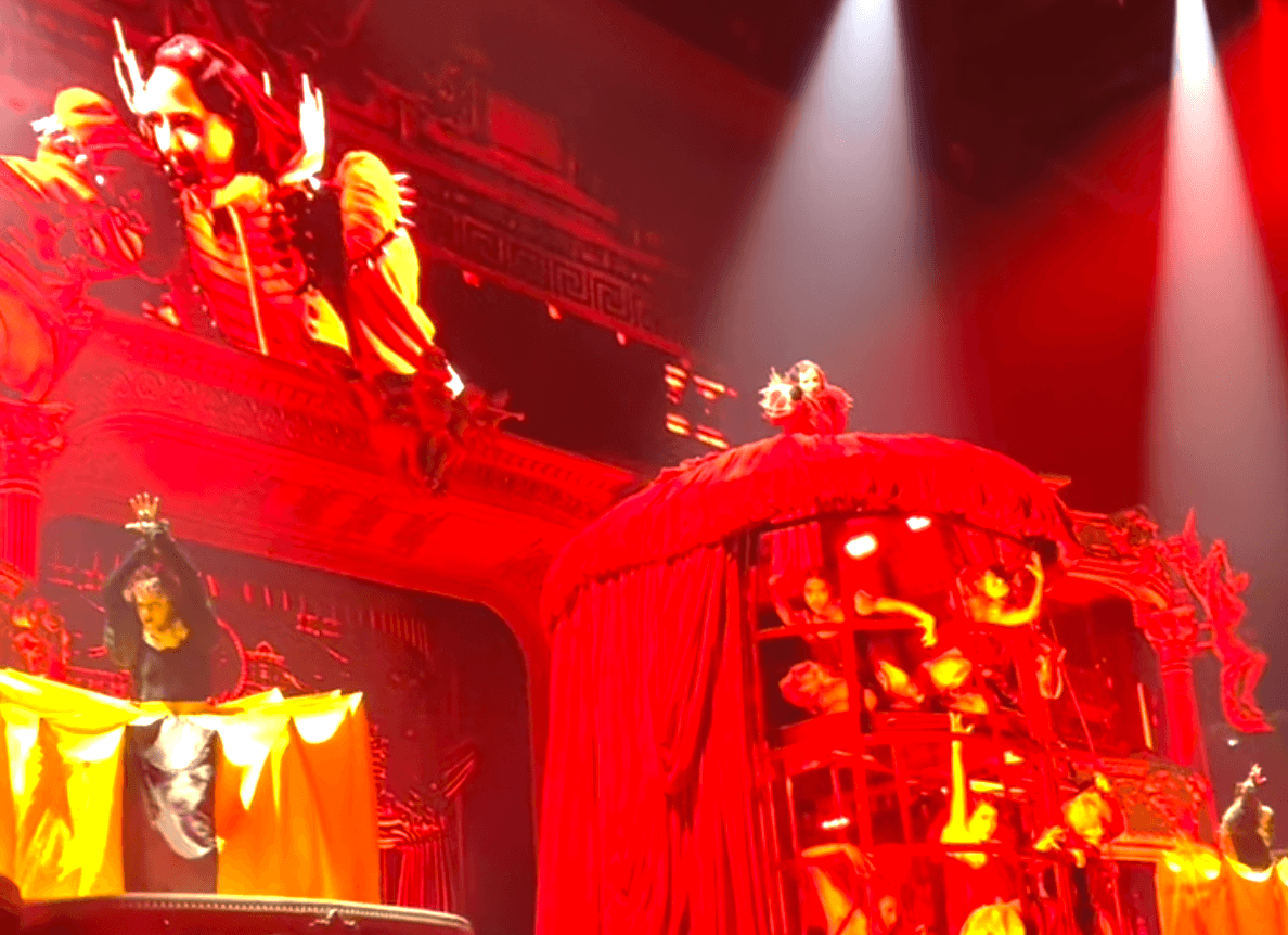 Lady Gaga on stage. She stands on top of a metal cage covered in a giant red dress, and points at the camera. There's a lot of people inside that cage