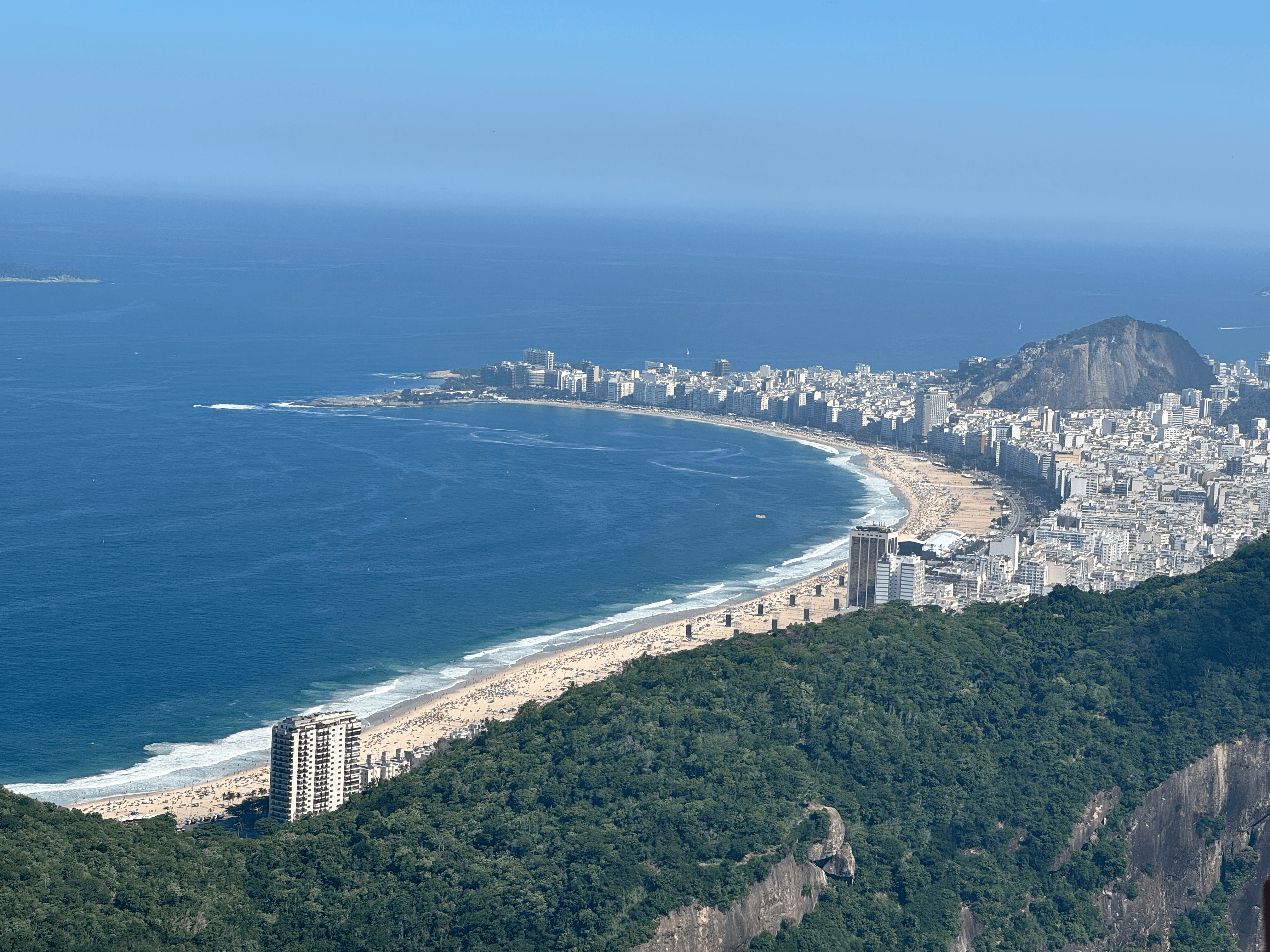 Picture of Copacabana beach seen from above. Weather is sunny, sky and see are blue. The beach is curved and surrounded by tall buildings, which are themselves surrounded by native atlantic forests.