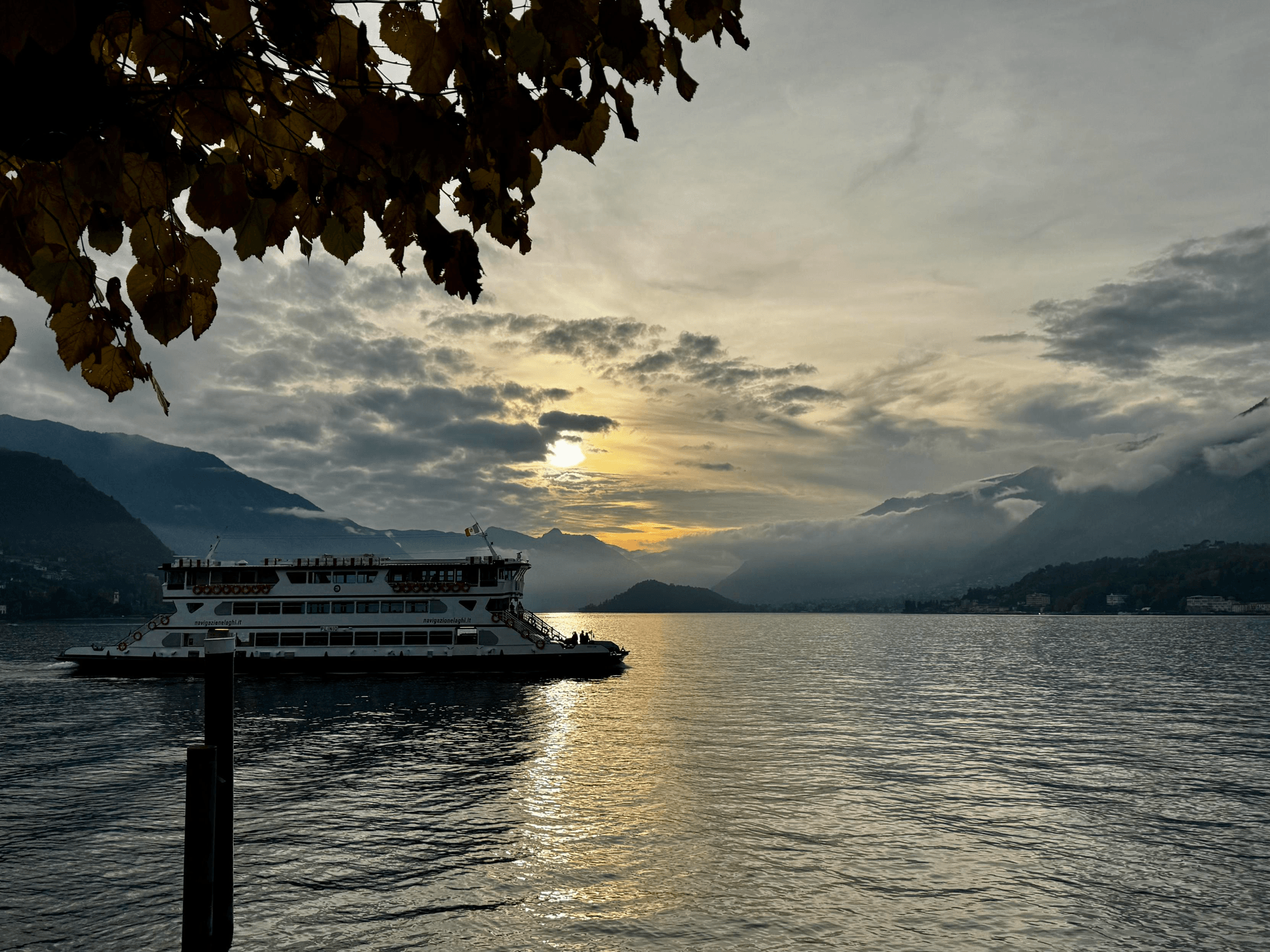 A large ferry crosses a calm lake at sunset, with soft golden light reflecting on the water. In the background, mist and clouds drift around dark mountain slopes, while autumn leaves frame the top of the image. The atmosphere is peaceful and slightly moody, evoking the quiet transition from day to evening.