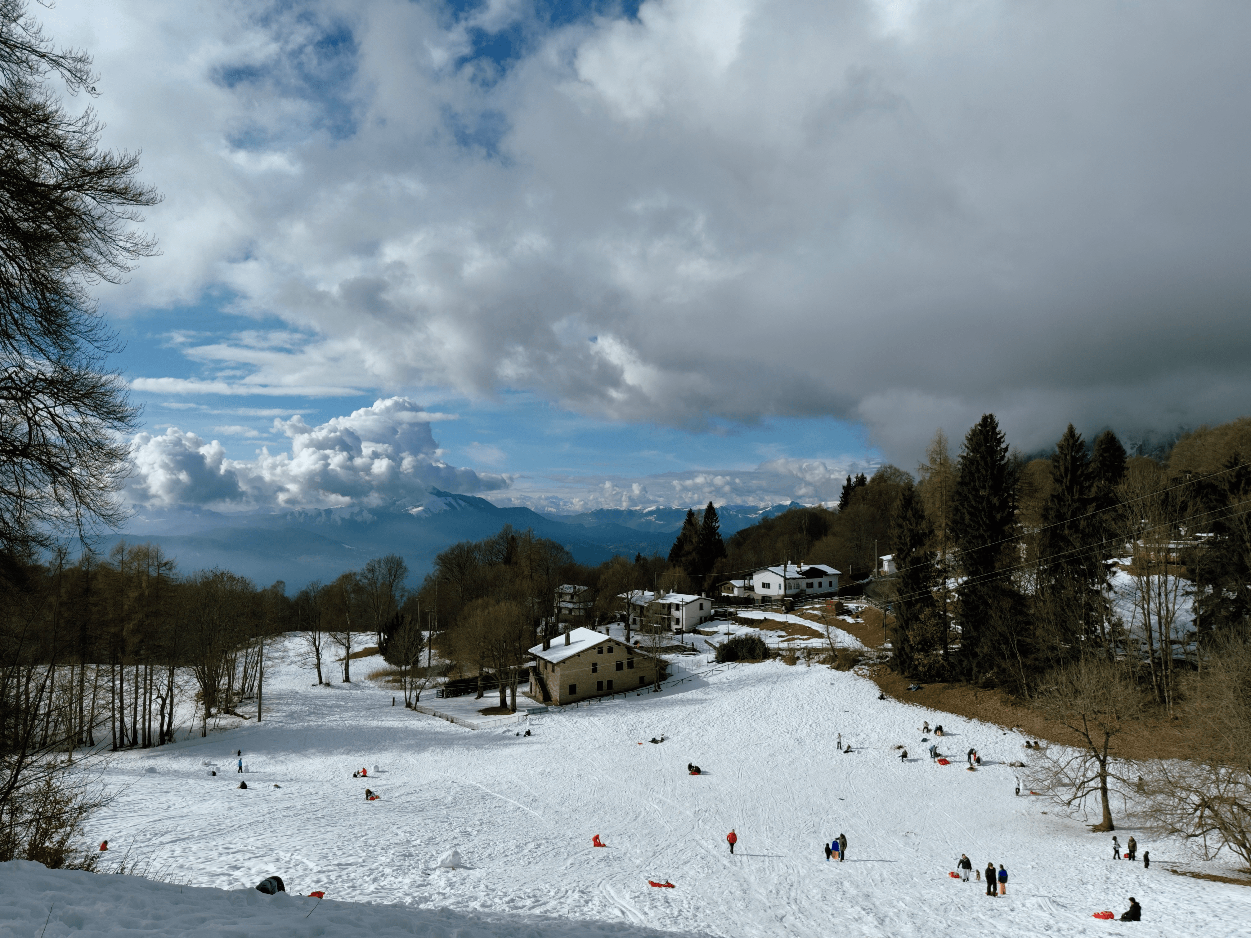 An open area full of snow and people playing on it. There's an alpine village a bit further ahead, and in the background there's the snow-capped Alps. The sky is partly cloudy.
